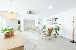 A spacious living area with a wooden dining table, white wicker chairs, and a large woven pendant light. There are several potted plants and a tall wooden sculpture. - Apartamentos Martalia Nerja (Torrox)