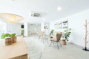A spacious living area with a wooden dining table, white wicker chairs, and a large woven pendant light. There are several potted plants and a tall wooden sculpture.