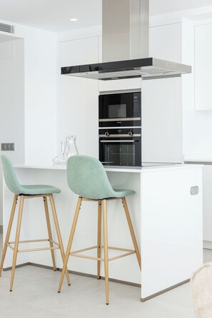 A modern kitchen with a built-in oven and microwave, two bar stools, and a white countertop. - Apartamentos Martalia Nerja (Torrox)