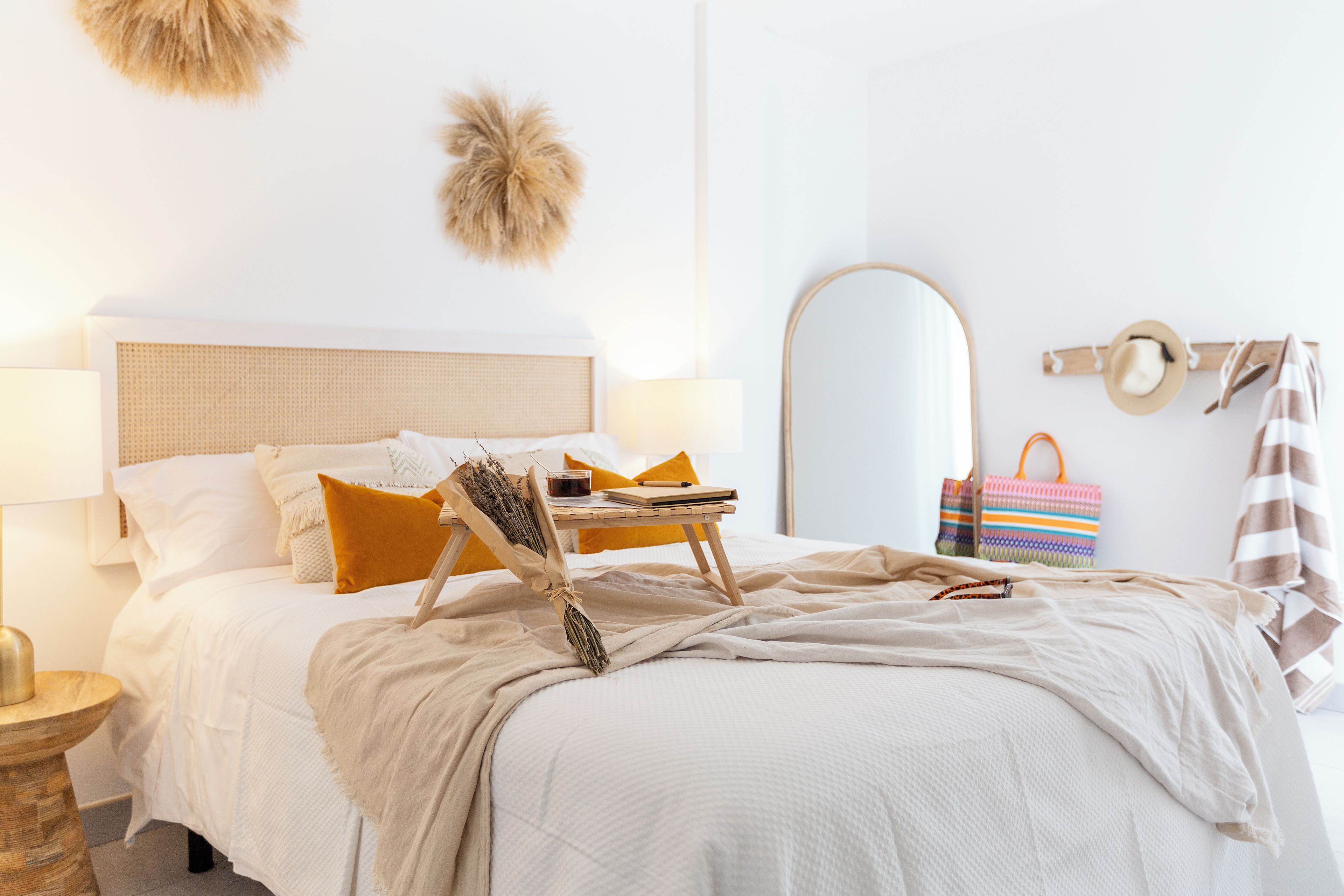 A neatly made bed with a headboard, a bedside table with a lamp, a mirror, and a wall-mounted hat rack.