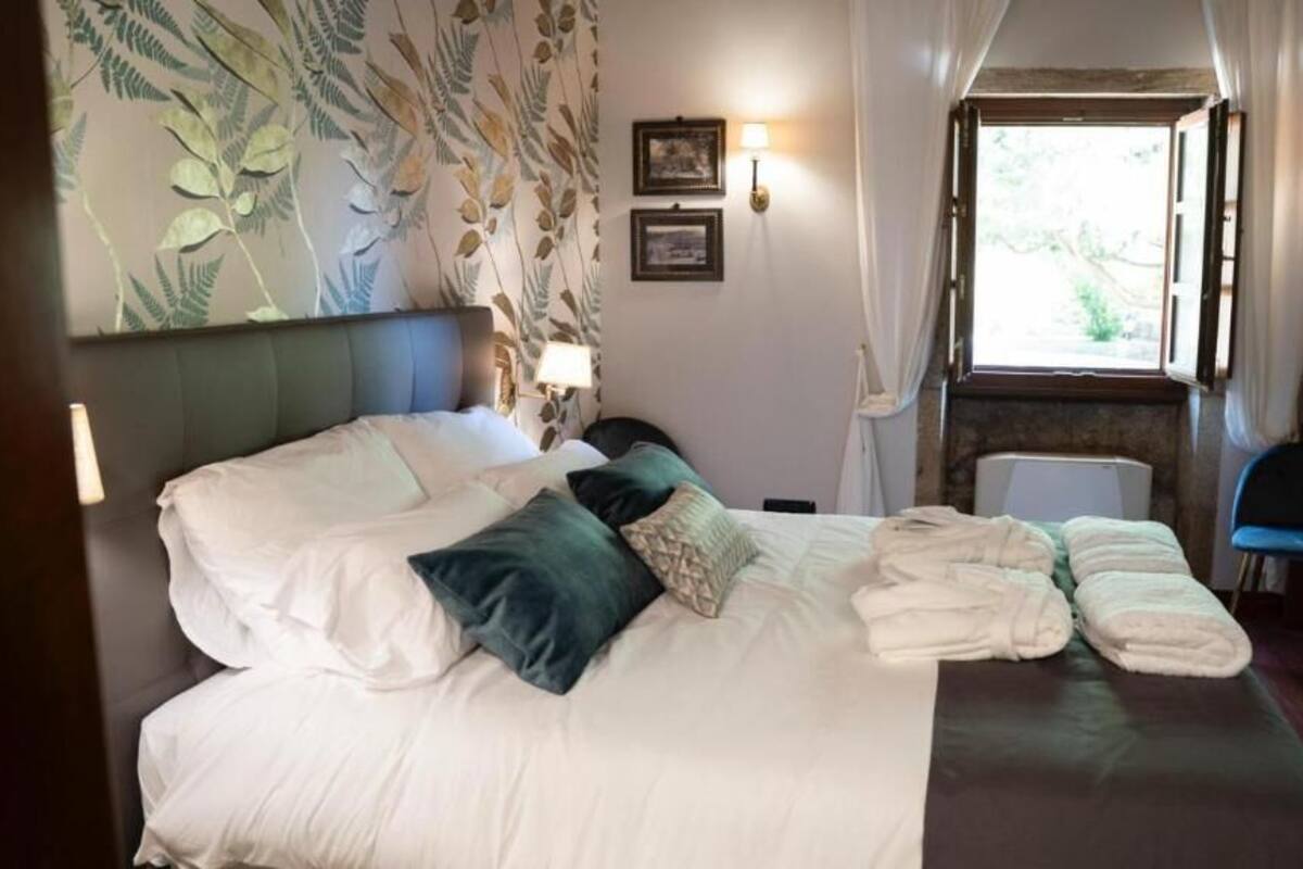 A hotel room with a large bed, two bedside lamps, a window with curtains, and a wall with a leafy pattern.