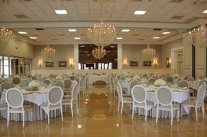 Un salón de banquetes con mesas redondas, sillas blancas, candelabros y cuadros en las paredes.
