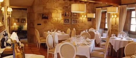 A restaurant with stone walls, wooden beams, and white tablecloths. Tables are set with wine glasses and plates.