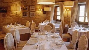 A restaurant with stone walls, wooden beams, and chandeliers. Tables are set with white tablecloths and checkered chairs. - Snö Pazo de Adran (Teo)