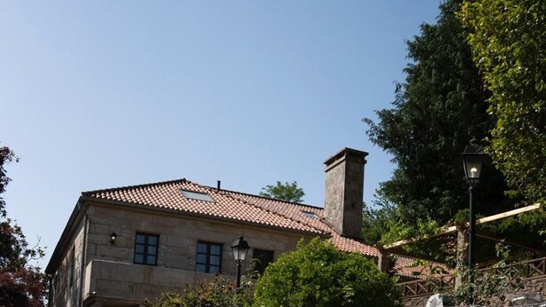 A swimming pool with clear blue water, a stone wall with a lifebuoy, and a building with a tiled roof in the background. - Snö Pazo de Adran (Teo)