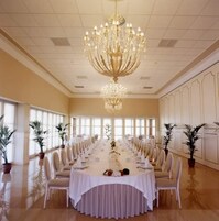 Un amplio salón de conferencias con una larga mesa dispuesta para un evento formal, rodeado de plantas en macetas y una gran lámpara de araña.