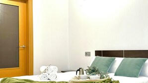 Una cama bien hecha con ropa de cama verde, una puerta de madera y una mesita de noche con una tetera y una planta.