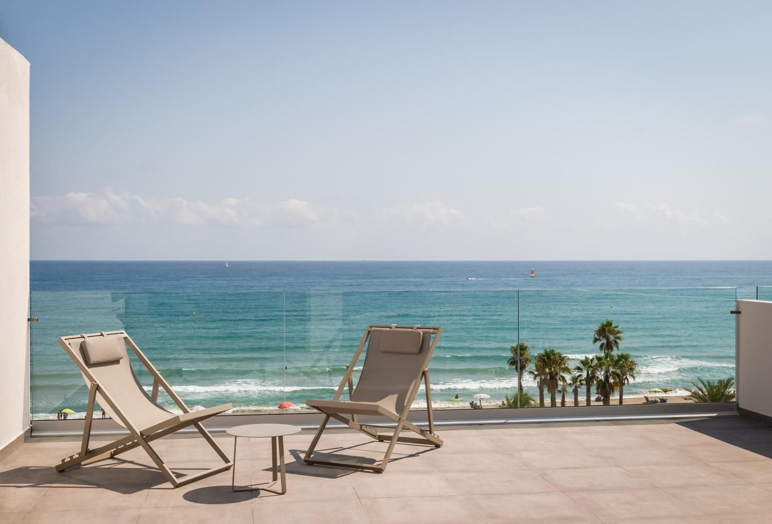 A balcony with two lounge chairs and a small table, overlooking a beach with palm trees and the ocean.