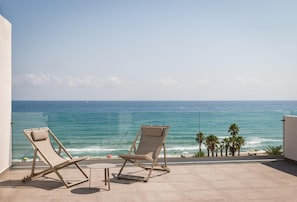 A balcony with two lounge chairs and a small table, overlooking a beach with palm trees and the ocean. - Estival Maramar (El Vendrell)