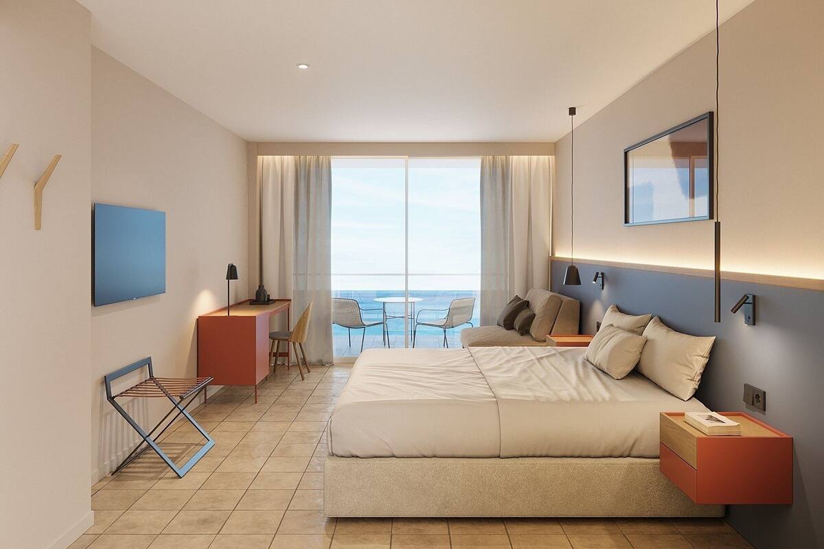 A modern hotel room with a large bed, a desk, and a balcony with a view of the sea.