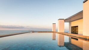 A serene infinity pool with a clear view of the ocean, a wooden deck, and a modern building with large pillars. - Estival Maramar (El Vendrell)