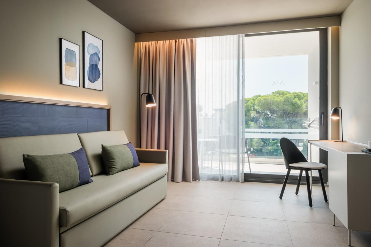 Een moderne hotelkamer met een bank, een bureau en een stoel. Twee ingelijste kunstwerken hangen aan de muur, een groot raam met gordijnen en uitzicht op groen buiten.