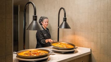 A chef preparing paella in a kitchen with two large pans on a counter, under two black pendant lights.