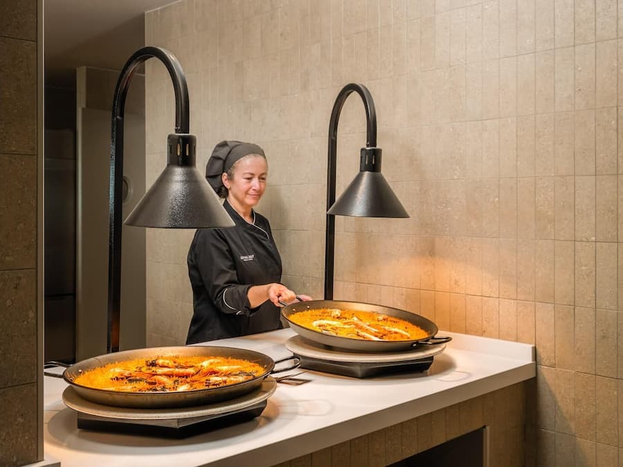 A chef preparing paella in a kitchen with two large pans on a counter, under two black pendant lights.