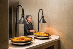 A chef preparing paella in a kitchen with two large pans on a counter, under two black pendant lights. - Estival Maramar (El Vendrell)