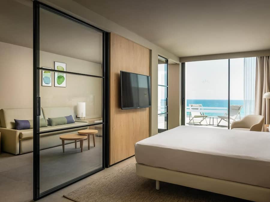 Habitación de hotel con una cama grande, televisión, sofá y balcón con vista al océano.
