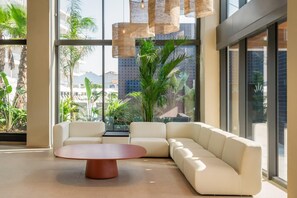 A modern lounge area with a sectional sofa, a round coffee table, and large windows offering a view of palm trees and buildings. - Estival Maramar (El Vendrell)
