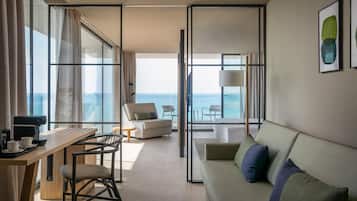 A modern hotel room with a large window offering a view of the sea, a desk with a coffee maker, a sofa, and a chair.