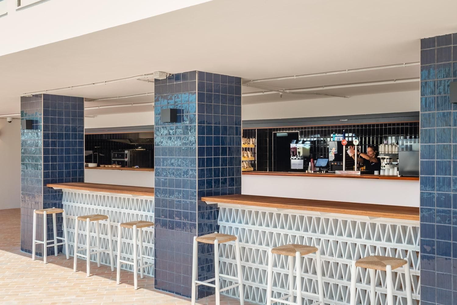 Zona de bar con taburetes altos y una barra, con columnas de azulejos azules y una máquina de café a la vista.