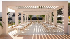 An outdoor patio with multiple tables and chairs, covered by a white pergola, and surrounded by greenery. - Estival Maramar (El Vendrell)