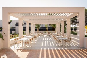 Um pátio externo com várias mesas e cadeiras, coberto por uma pérgola branca e rodeado por vegetação.