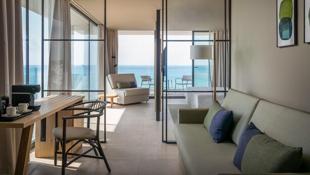 A modern hotel room with a large window offering a view of the sea, a desk with a coffee maker, a sofa, and a chair. - Estival Maramar (El Vendrell)