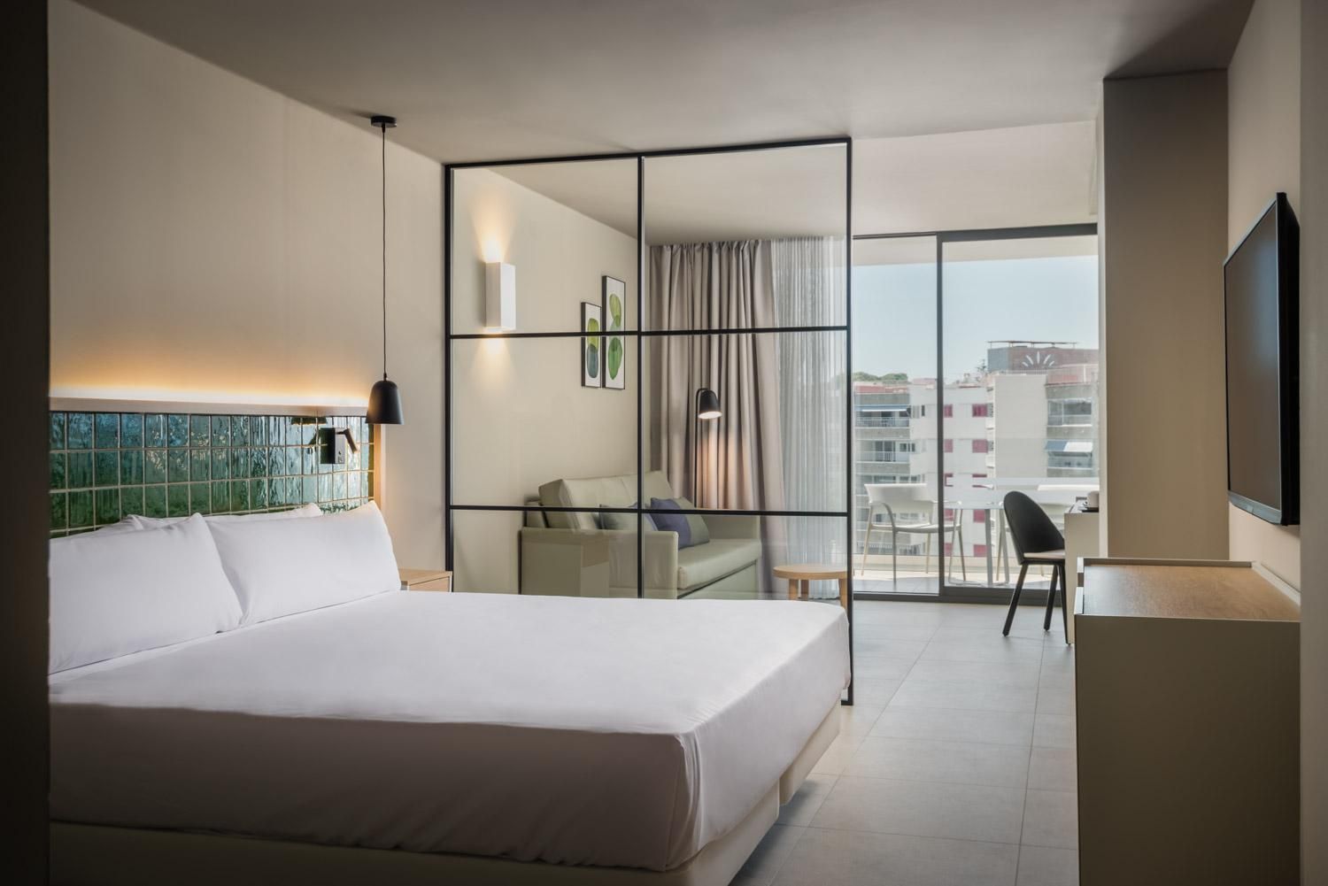 A hotel room with a large bed, a desk, a chair, and a balcony with a view of the city.