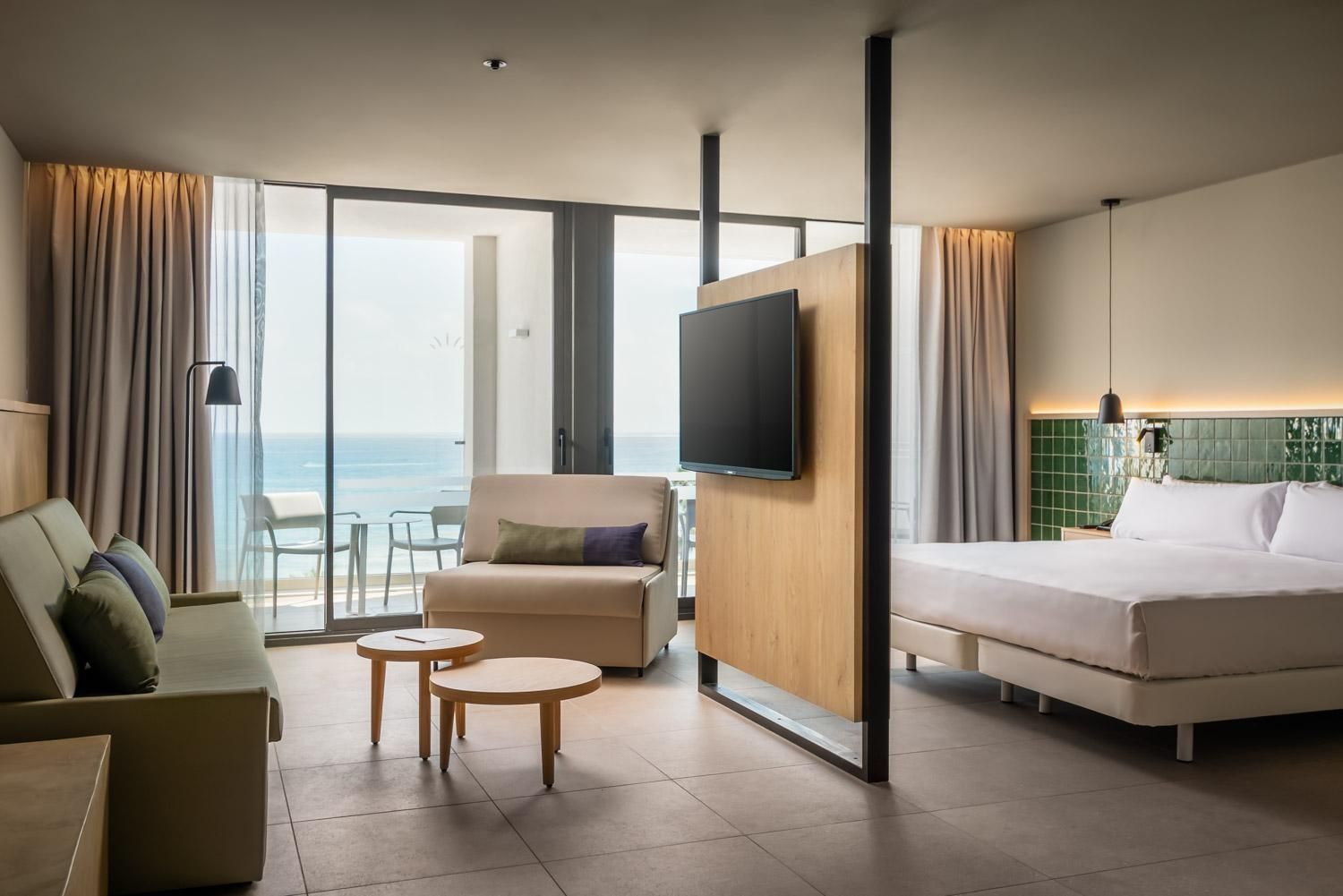 A hotel room with a bed, a sofa, a TV, a small table, and a balcony with a view of the sea.