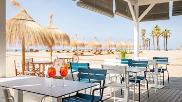 Un restaurante junto a la playa con terraza al aire libre, sombrillas de paja y palmeras.