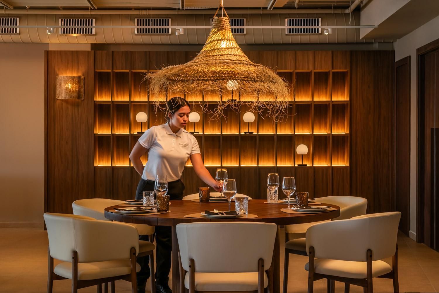 Un ambiente de restaurante con pared de madera, una araña colgante y un camarero atendiendo una mesa.