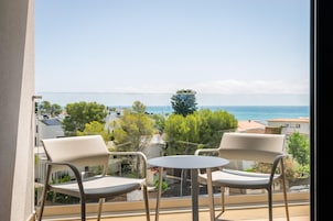 A balcony with two chairs and a small table, overlooking a coastal view with buildings and trees.