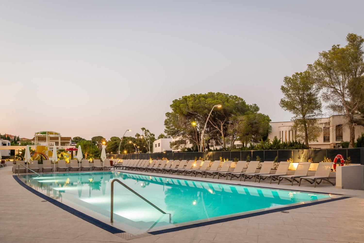Una piscina con sillas de descanso y sombrillas, rodeada de árboles y edificios.