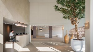 A modern reception area with a receptionist, a reception desk, a large potted tree, and a seating area with sofas. - Estival Maramar (El Vendrell)