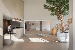 A modern reception area with a receptionist, a reception desk, a large potted tree, and a seating area with sofas. - Estival Maramar (El Vendrell)