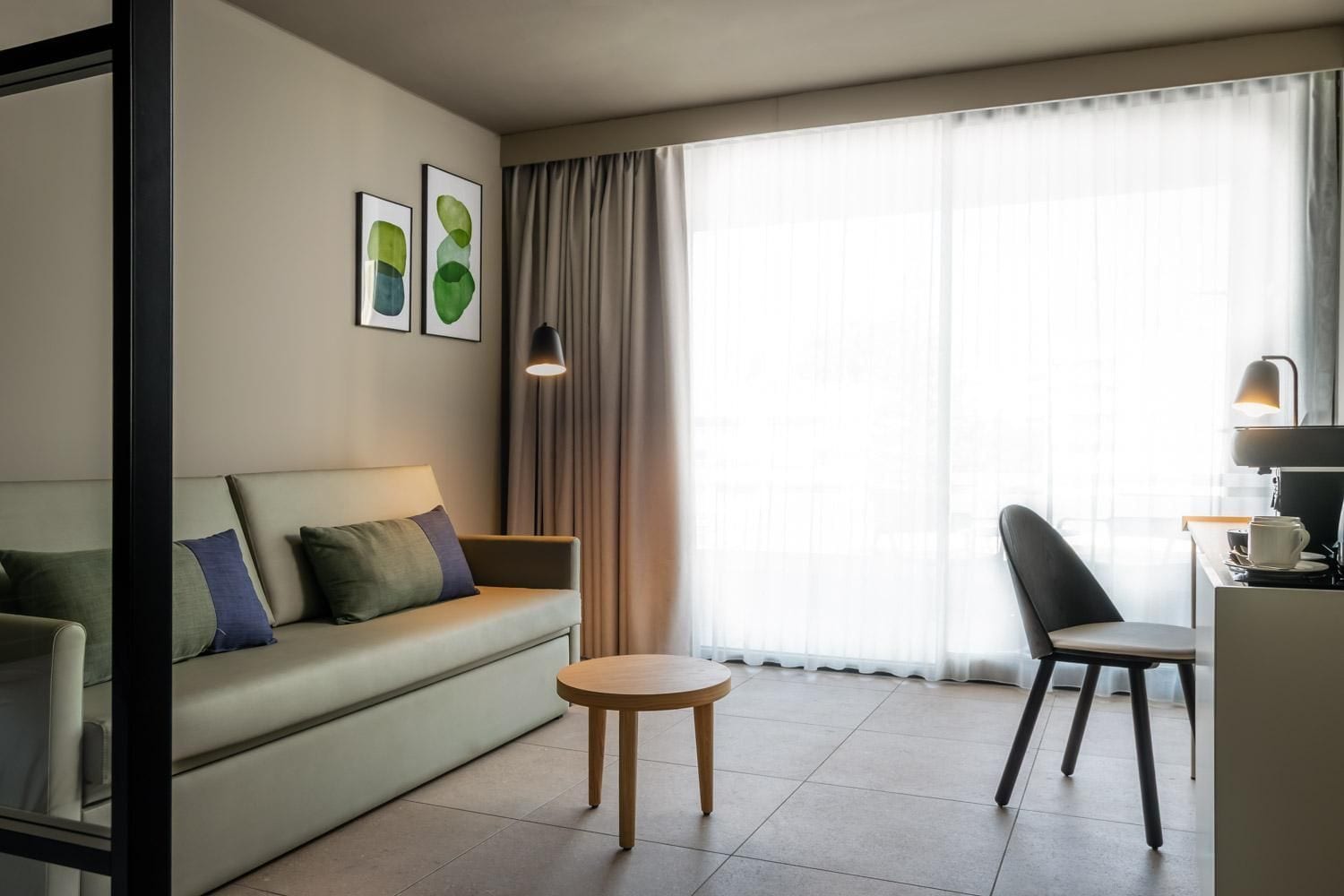 Una habitación de hotel moderna con sofá, una pequeña mesa redonda, una silla, un escritorio con lámpara y un gran ventanal con cortinas.