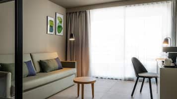 Una habitación de hotel moderna con sofá, una pequeña mesa redonda, una silla, un escritorio con lámpara y un gran ventanal con cortinas.