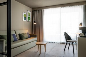 A modern hotel room with a sofa, a small round table, a chair, a desk with a lamp, and a large window with curtains. - Estival Maramar (El Vendrell)