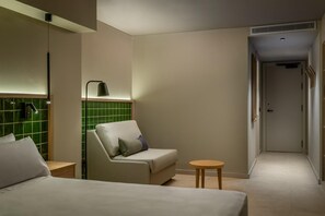 Habitación de hotel con cama, una silla, una mesita pequeña y un muro con azulejos verdes.