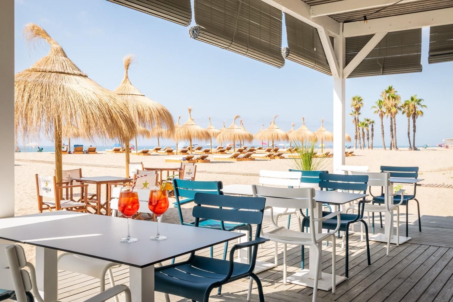 Un restaurante junto a la playa con terraza al aire libre, sombrillas de paja y palmeras.