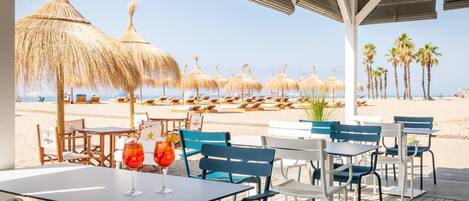 A beachside restaurant with outdoor seating overlooking the beach, thatched umbrellas, and palm trees.
