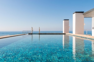 Uma piscina infinita azul cristalina com vista para o oceano, pilares brancos e uma área coberta.