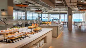 A buffet area with various food options, a seating area with tables and chairs, and large windows offering a view of the ocean. - Estival Maramar (El Vendrell)