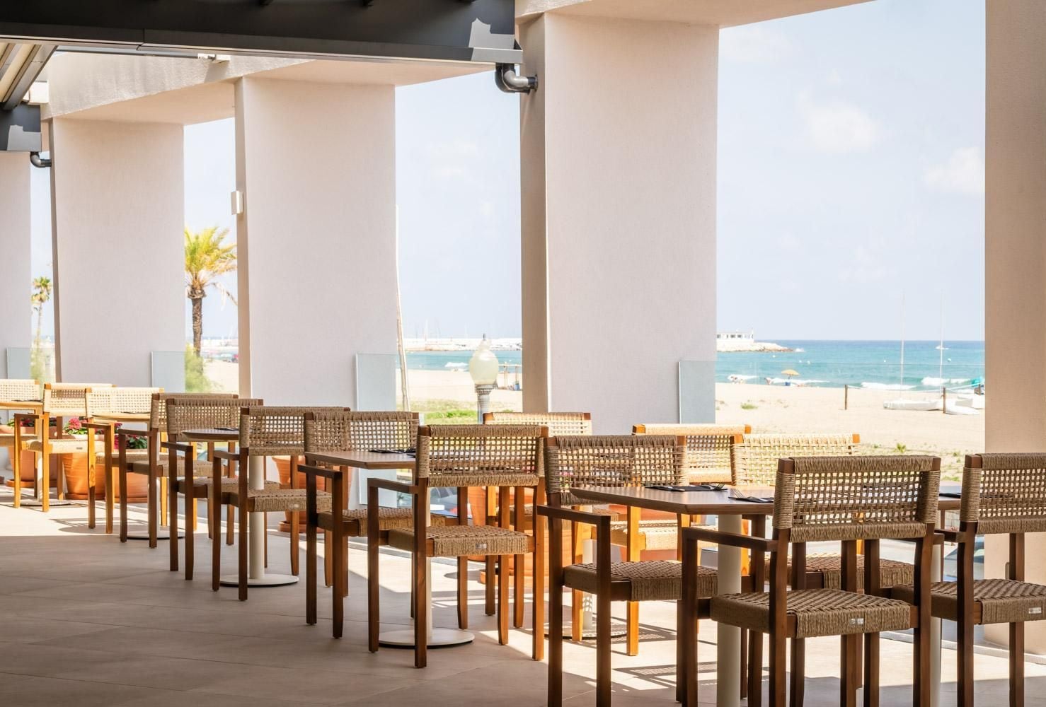 An outdoor seating area with wicker chairs and tables, overlooking a beach and the sea.