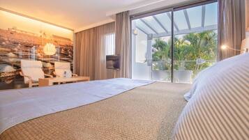 Habitación de hotel con una cama grande, una zona de estar con sillas, un televisor y un balcón con vistas a palmeras.