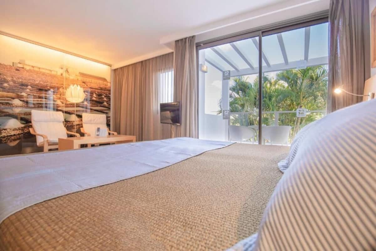 Une chambre d’hôtel dotée d’un grand lit, d’un coin salon avec des fauteuils, d’une télévision et d’un balcon offrant une vue sur les palmiers.