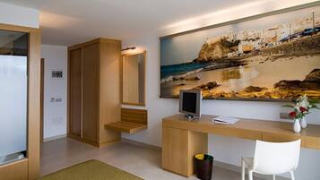 Habitació d'hotel moderna amb escriptori de fusta, un ordinador, cadira blanca, paperera petita i un ampli quadre de platja a la paret.