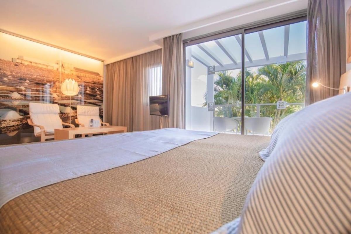 A hotel room with a large bed, a seating area with chairs, a TV, and a balcony with a view of palm trees.