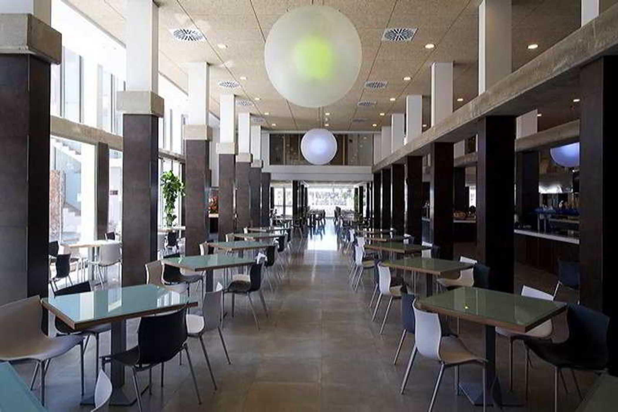 Un espacio interior amplio con varias mesas y sillas, ventanas grandes y un techo alto con una lámpara central.