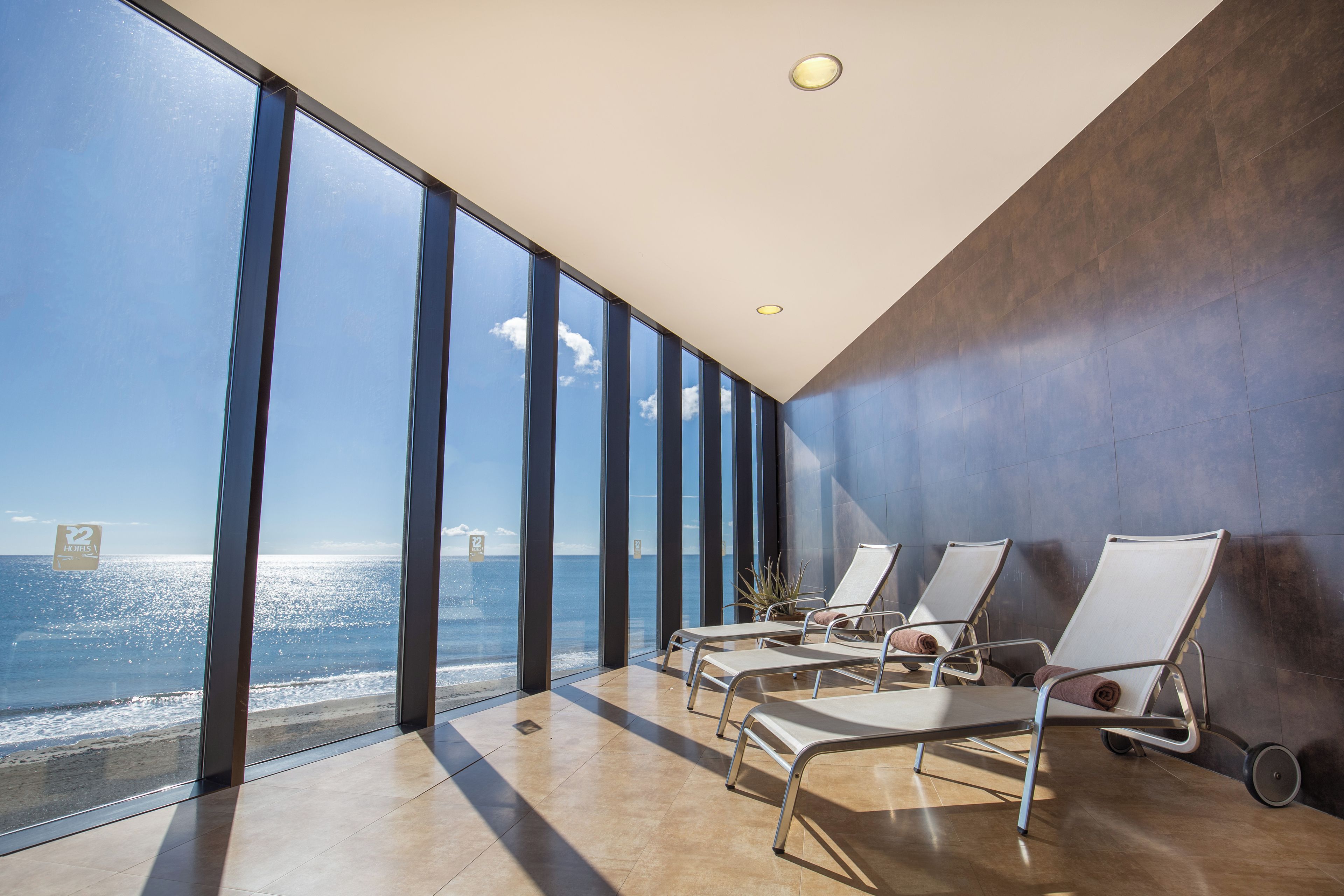 Habitación de hotel moderna con ventanas amplias que ofrecen vista al mar, una tumbona y una planta en maceta.