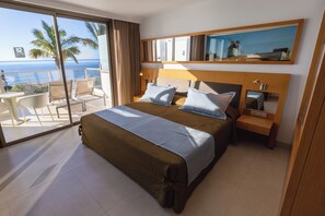 Une chambre d’hôtel avec un grand lit, une table de chevet, un bureau et une chaise. Une grande fenêtre offre une vue sur les palmiers et l’océan.